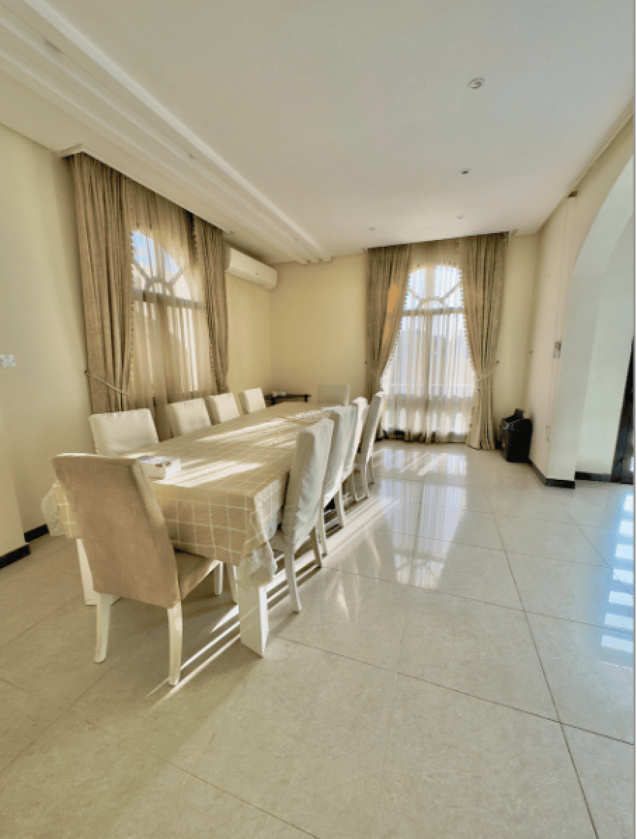 A dining room with a long table, beige tablecloth, and multiple white chairs. Sunlight streams through tall curtains on the windows.